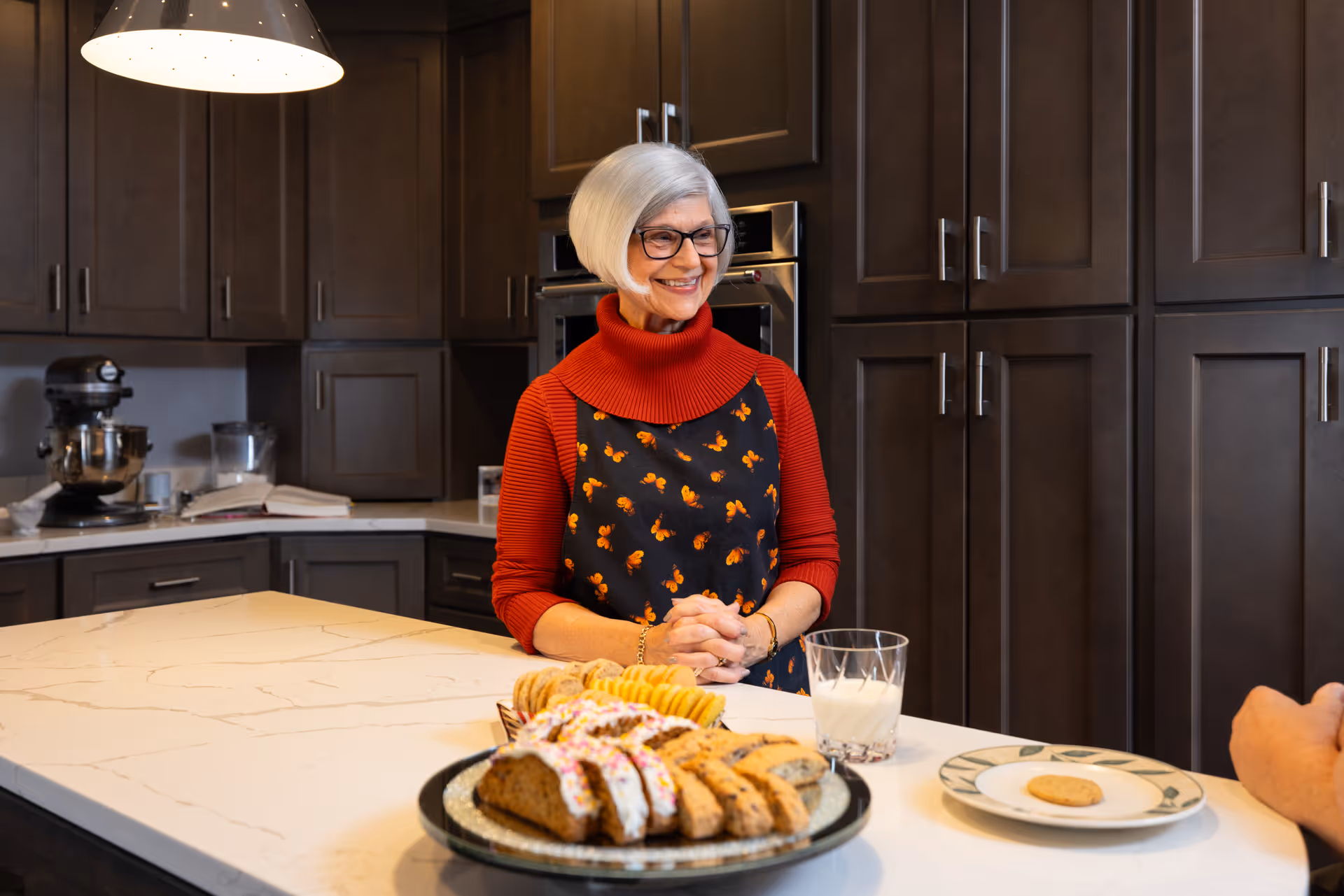 A smiling older woman in an apron stands behind a kitchen island with a plate of pastries and a glass of milk.
