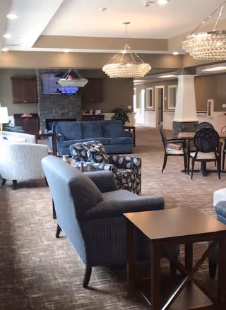 Spacious senior living common area with upholstered sofas and chairs, chandeliers, and a TV over a stone fireplace.