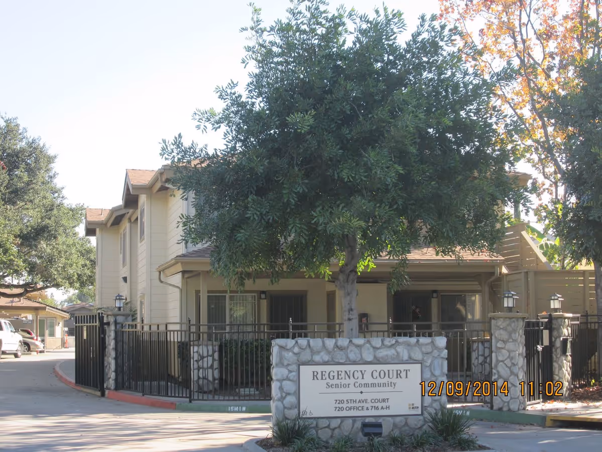 Exterior view of Regency Court Senior Community showing a gated entrance with a stone sign and a large tree in front. The building is two stories with beige siding and a brown roof. There are some cars parked to the left and trees surrounding the area.