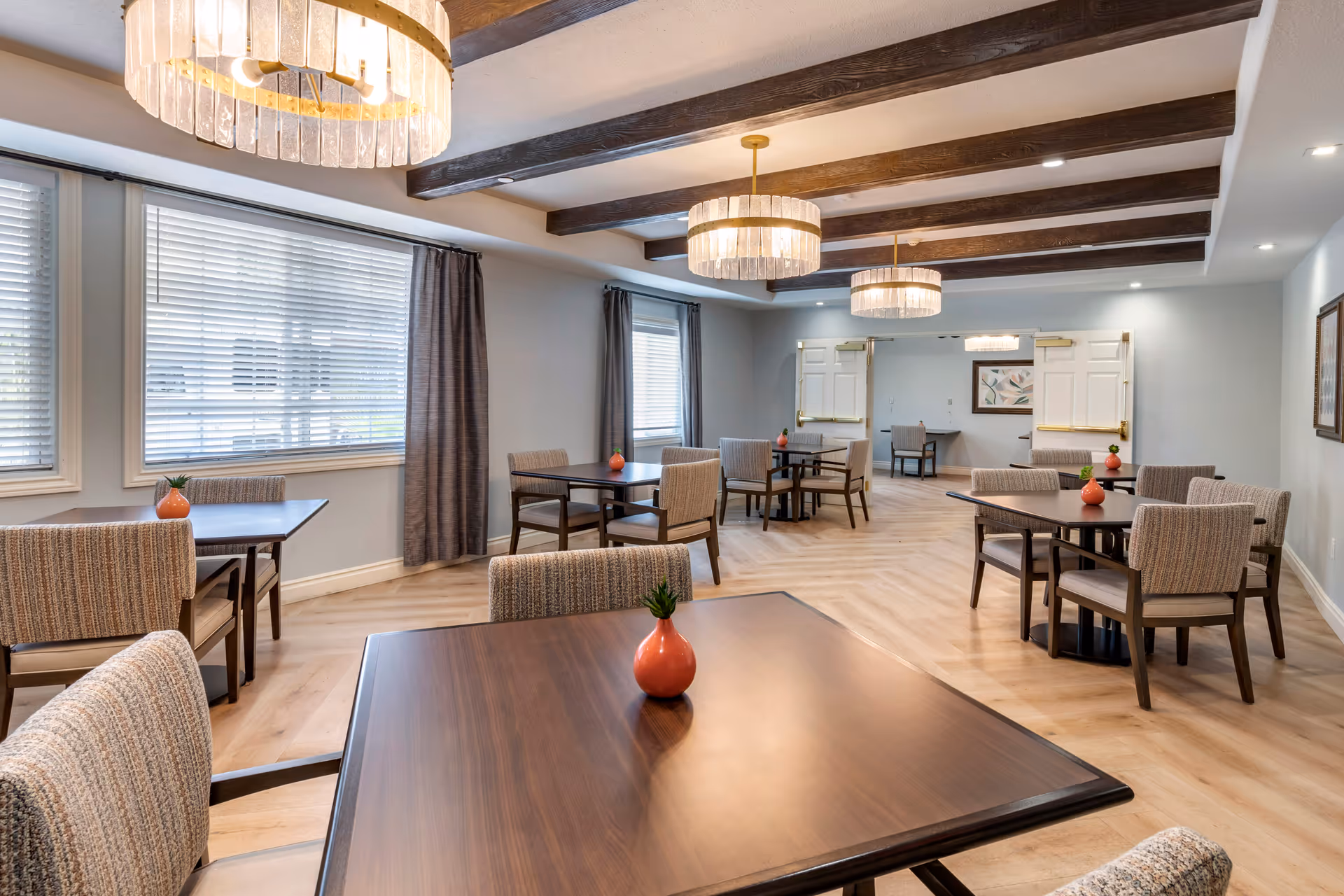 A bright and spacious dining room with several wooden tables and cushioned chairs arranged neatly. Each table has a small decorative orange vase with a green plant. The room features large windows with blinds and curtains, wooden ceiling beams, and three modern chandeliers providing warm lighting. The floor is light wood, and the walls are painted a soft gray with framed artwork.