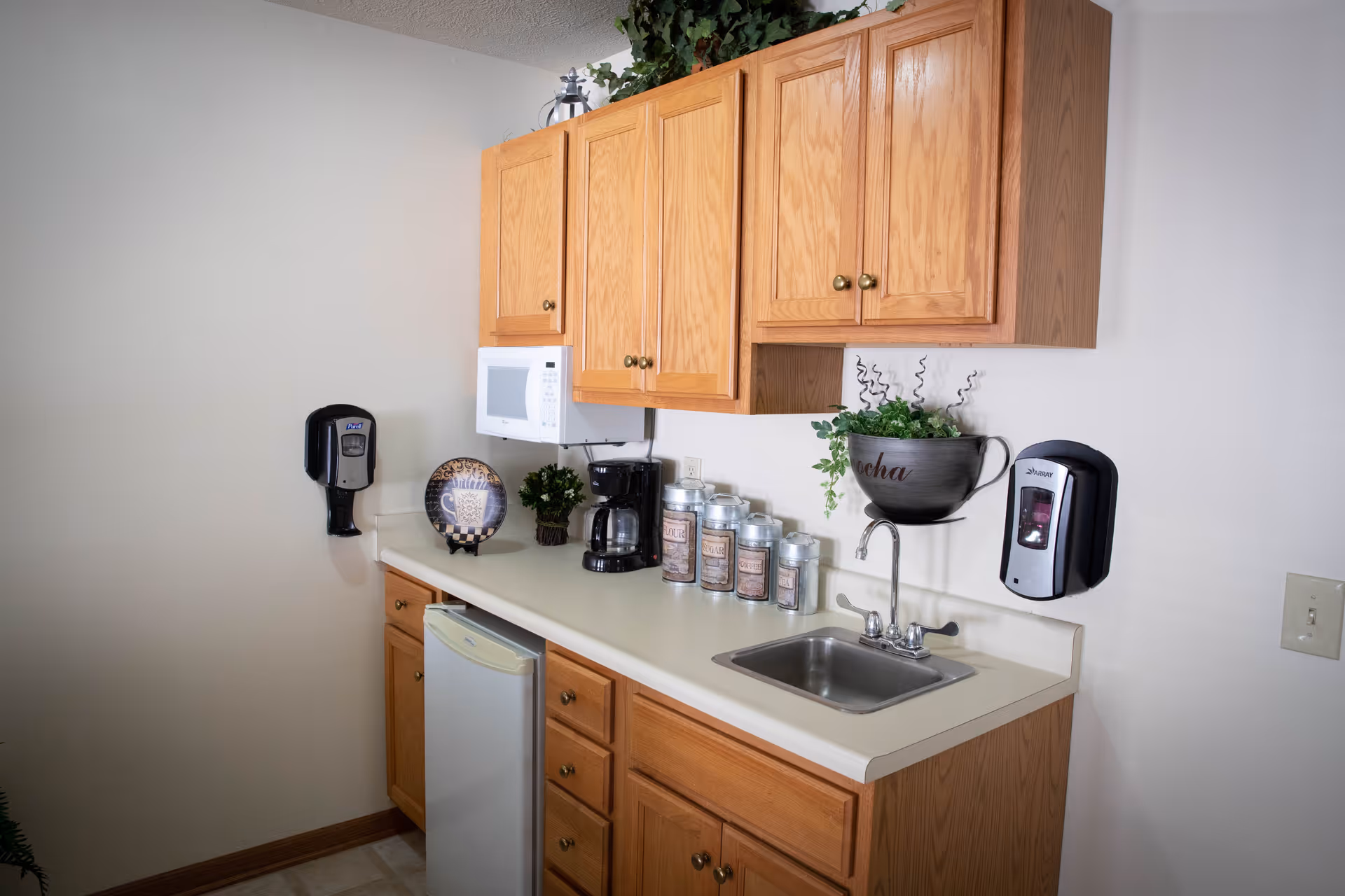 A small kitchenette area with wooden cabinets, a white countertop, a small sink, a coffee maker, a microwave, a mini refrigerator, and several labeled canisters. There are two wall-mounted dispensers and a decorative plant in a large cup-shaped container labeled 'Mocha'.