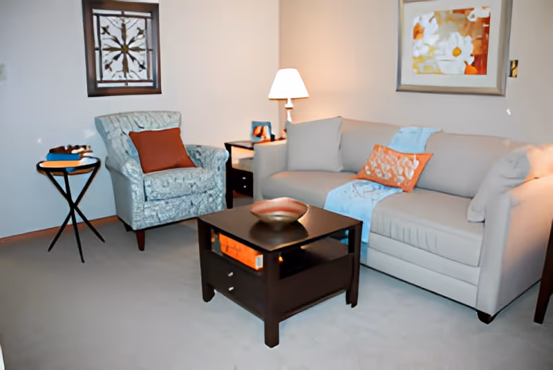 A living room with a beige sofa, patterned armchair, dark wood coffee table, side table with a lamp, and wall art.