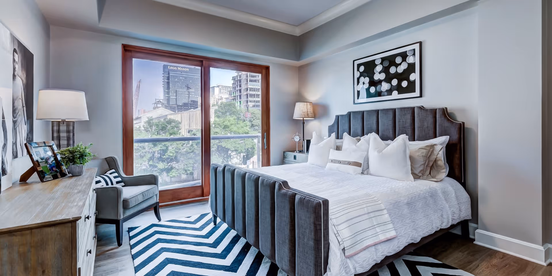 A modern bedroom with a large upholstered bed featuring multiple white pillows and a white bedspread. There is a wooden dresser with a lamp, framed photos, and a plant on top. A gray armchair with a striped pillow sits next to the dresser. The room has a large window with a wooden frame, showing an urban cityscape outside. The floor has a black and white chevron patterned rug, and there is a framed abstract artwork on the wall above the bed.