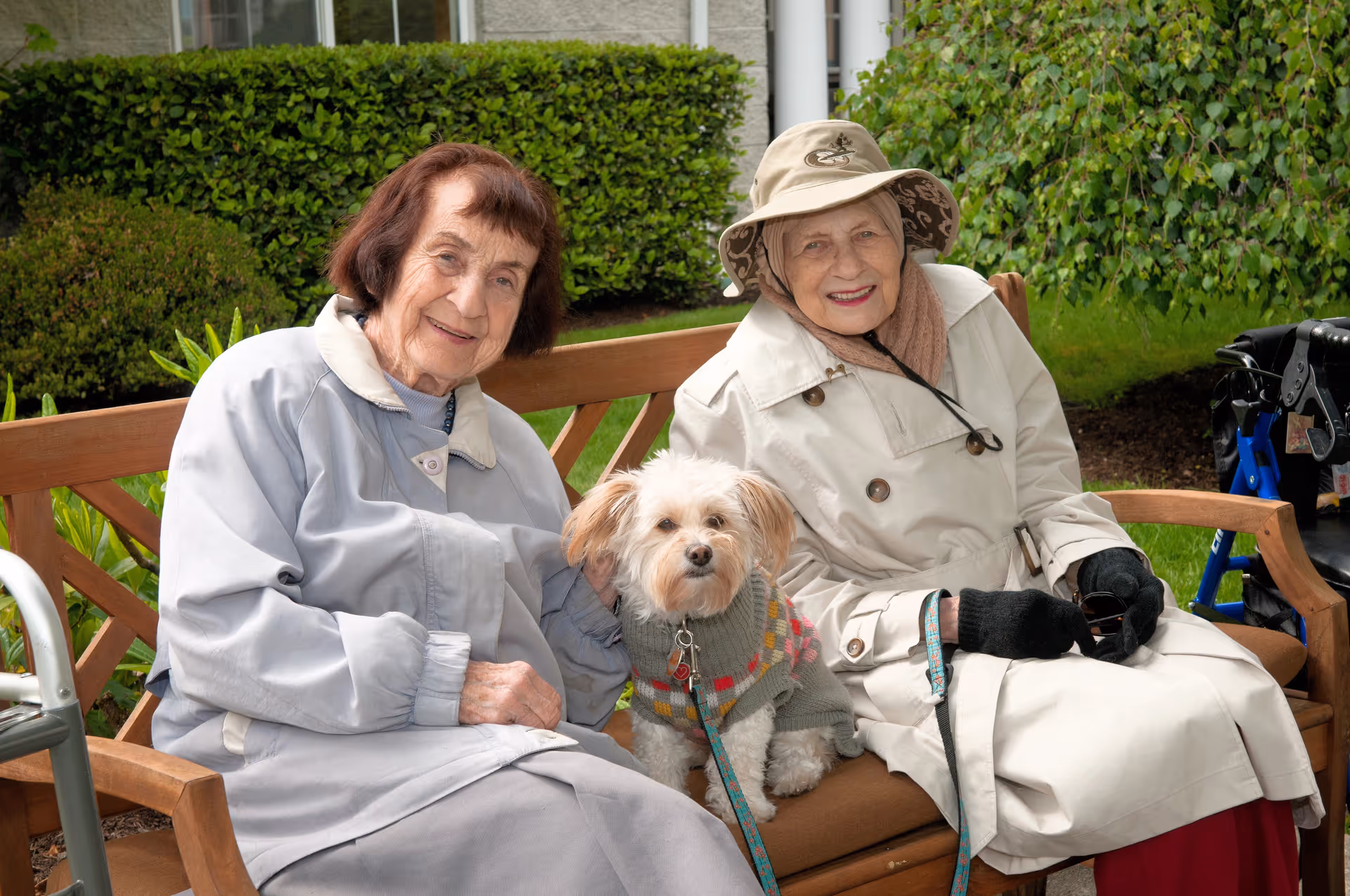 Two elderly women sitting on a wooden bench outdoors with a small dog wearing a sweater between them. One woman is wearing a light gray coat and the other is wearing a beige trench coat, hat, and black gloves. There is greenery and bushes in the background.