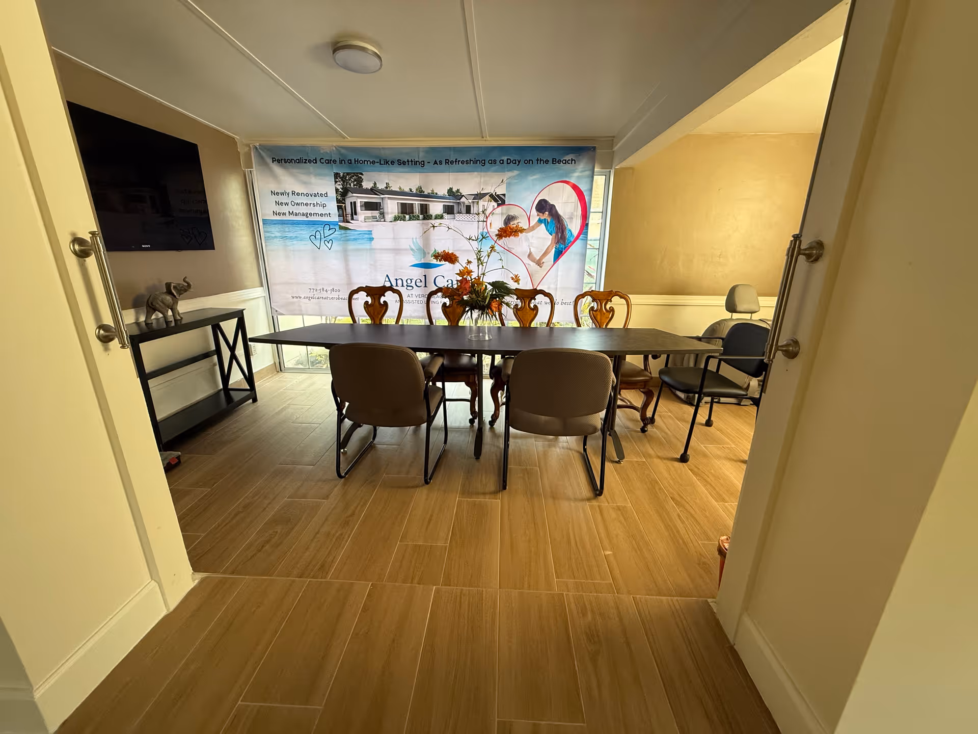 Interior view of a dining room with a rectangular table surrounded by chairs. A decorative flower arrangement is placed on the table. A large banner on the back wall displays information about Angel Care At Vero Beach, including an image of a woman and a house by the beach. The room has light-colored tiled flooring and beige walls. A TV is mounted on the left wall above a console table with a small elephant figurine.