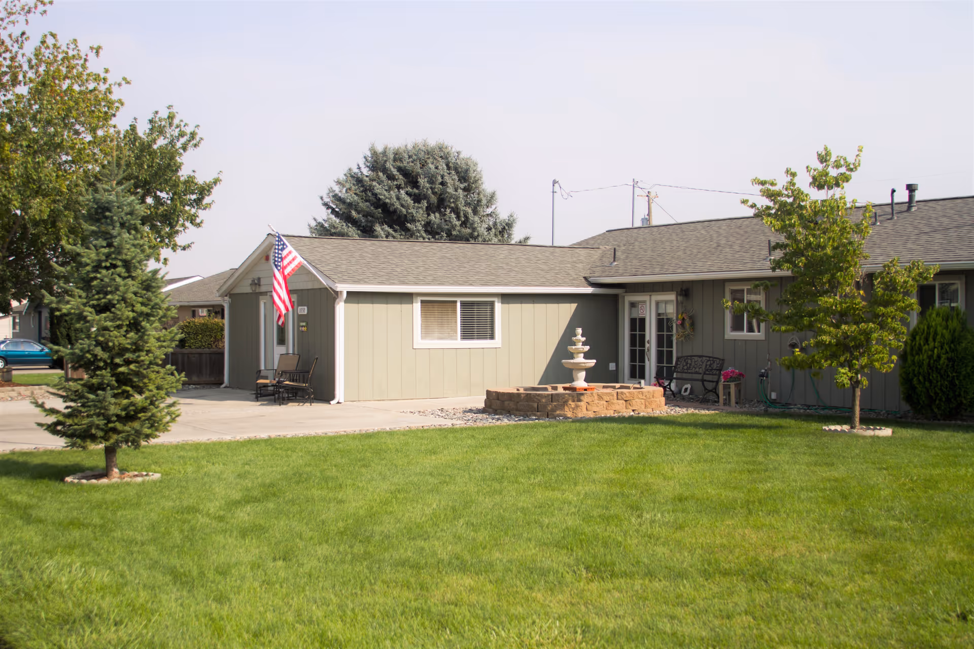Exterior view of a single-story residential care facility building with a well-maintained green lawn, two small trees, an American flag mounted near the entrance, outdoor seating, and a decorative fountain in front of the building.