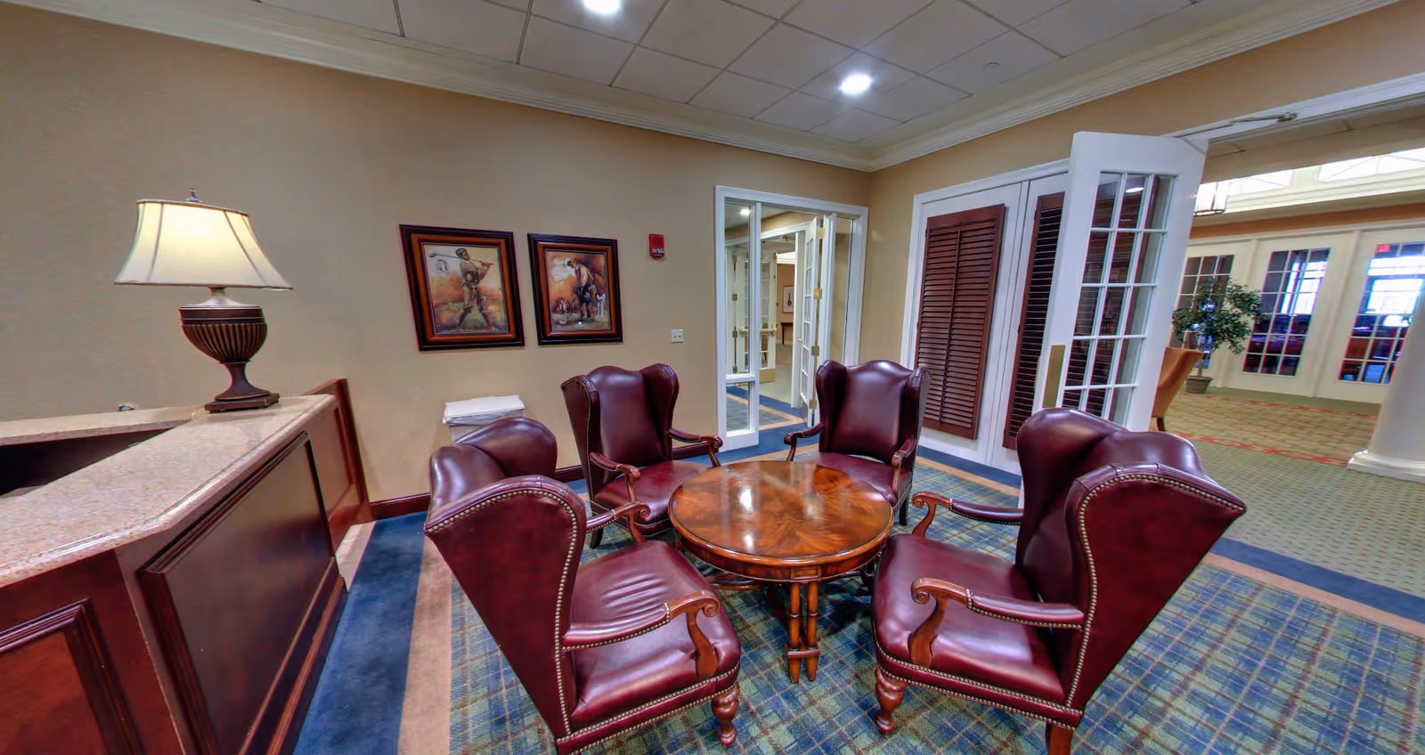 A cozy seating area in a senior living facility with five burgundy leather armchairs arranged around a round wooden table. The room has beige walls, a patterned carpet, two framed paintings on the wall, a table lamp on a wooden counter, and glass doors leading to another area with more seating and plants.