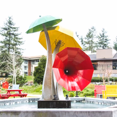 Outdoor sculpture featuring three large, colorful umbrella-like structures in green, yellow, and red, positioned in a water fountain with benches and a building in the background surrounded by trees and greenery.