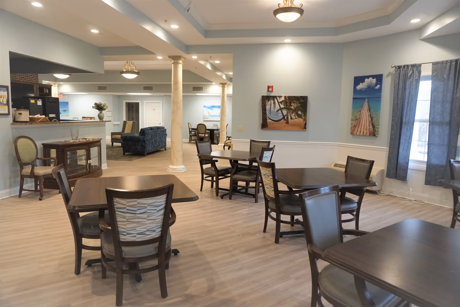 Interior view of a senior living facility dining area with several dark wood tables and chairs. The walls are painted light blue with white wainscoting, and there are two large paintings depicting beach scenes. The room has a window with blue curtains and recessed lighting in the ceiling. In the background, there is a lounge area with sofas and armchairs, and a counter area with a small cabinet and chair nearby.