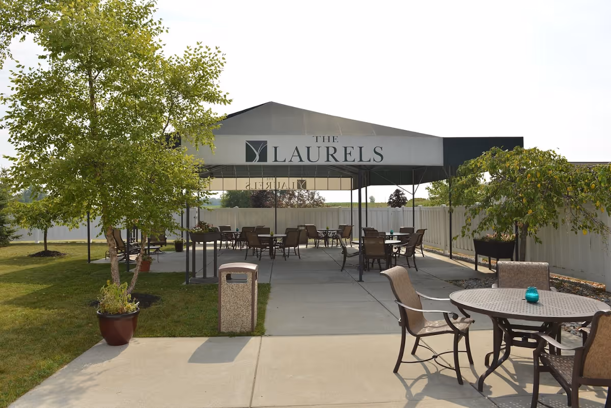 Outdoor seating area at The Laurels of Carson City featuring multiple tables and chairs under a large canopy with the facility name displayed on it. The area is surrounded by a white fence, with trees and potted plants nearby on a sunny day.
