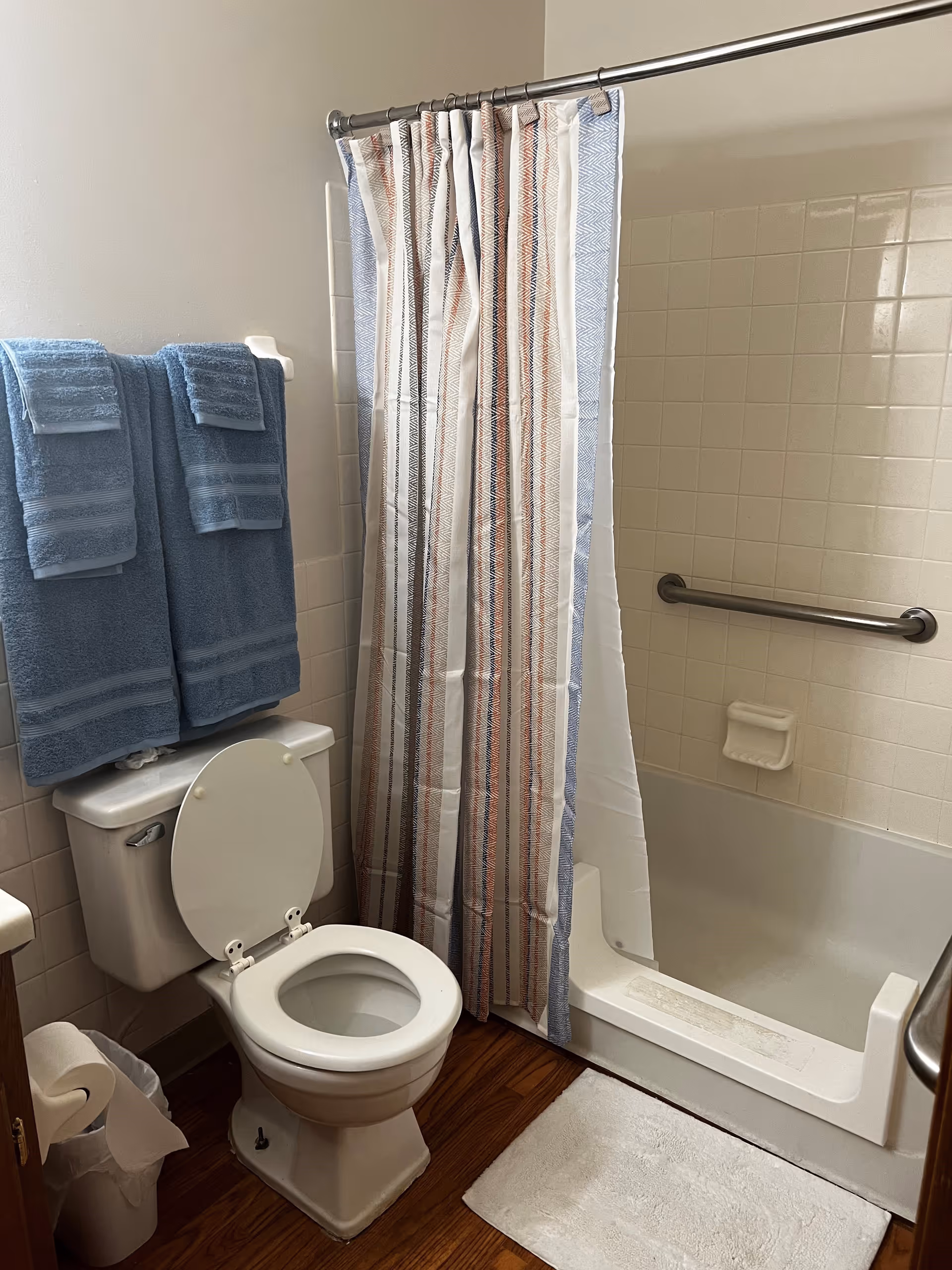 A bathroom with a white toilet with the lid open, two blue towels hanging on a rack above the toilet, a shower with a striped shower curtain, a white bath mat on the wooden floor, and a grab bar on the tiled wall inside the shower.