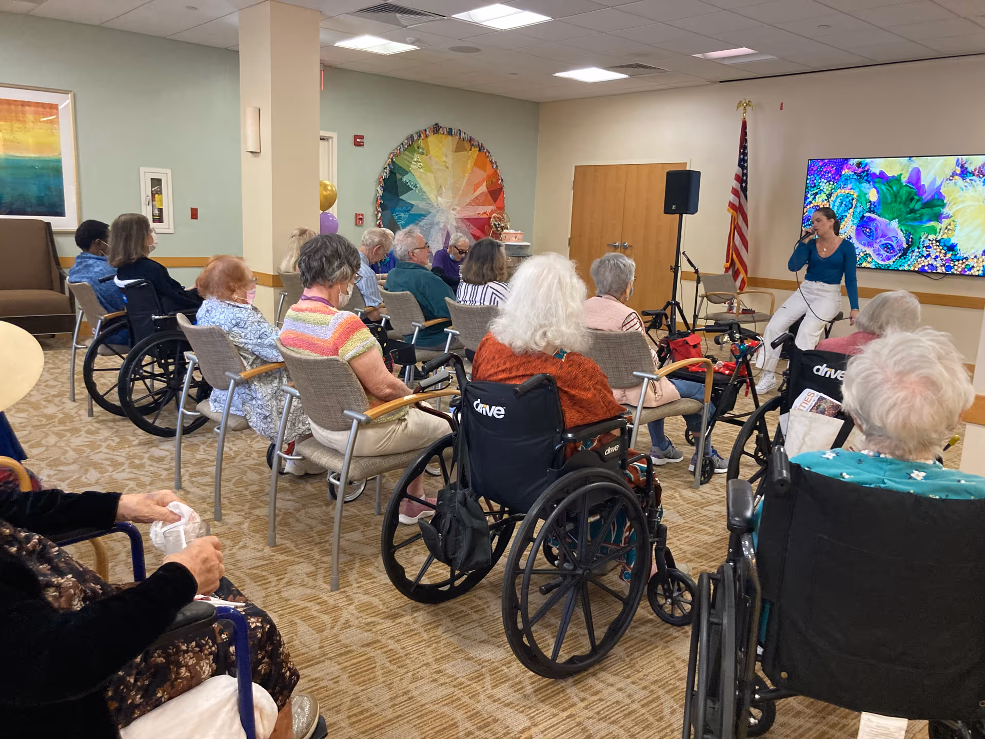 A group of elderly people, some in wheelchairs, seated in a room facing a woman who is speaking or singing into a microphone. The room has beige walls, a colorful circular wall decoration, an American flag, and a large screen displaying vibrant colors.