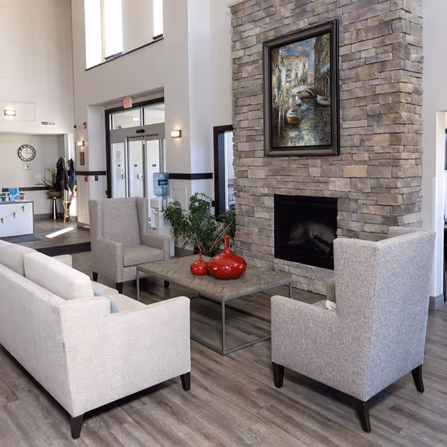 Lobby seating area in a senior living facility featuring sofas and armchairs arranged around a coffee table and a stone fireplace.