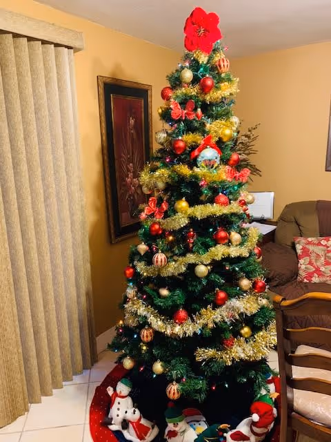 A decorated Christmas tree with red, gold, and white ornaments, gold tinsel garlands, and a red flower topper in a cozy living room. At the base of the tree are festive holiday figurines including snowmen and Santa Claus. The room has beige walls, a framed floral painting, vertical blinds on a window, and a brown sofa with patterned pillows.