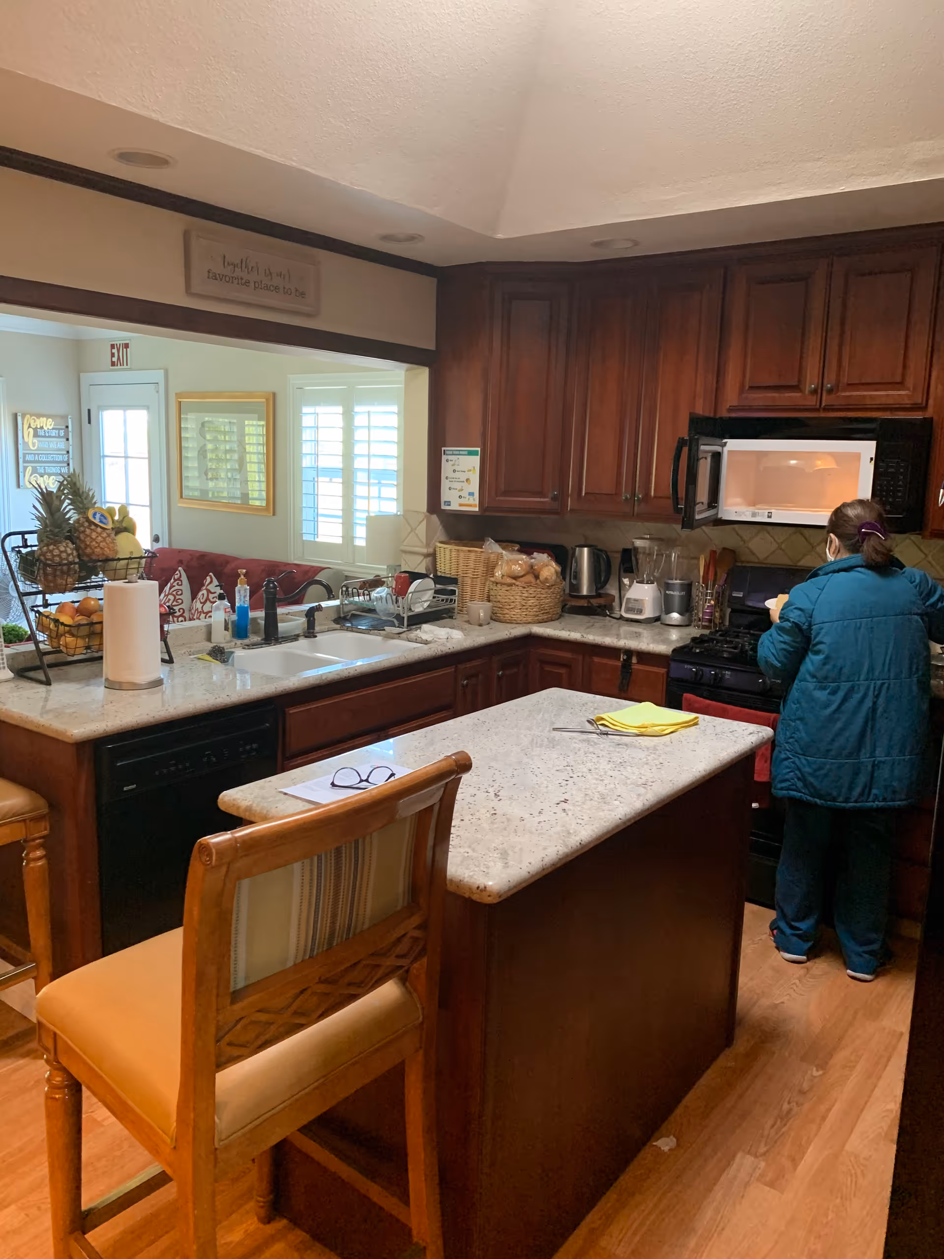 A kitchen with wooden cabinets, a marble countertop island with a chair, and various kitchen appliances including a microwave, blender, and kettle. A person wearing a teal jacket is standing near the stove. In the background, there is a living room area with a red couch and windows with white shutters.