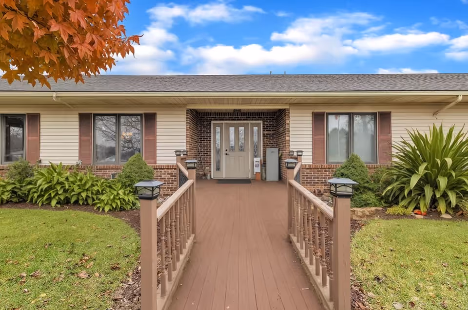 Front exterior view of a single-story building with a brown wooden ramp leading to a beige door with glass panels. The building has beige siding with brick accents and brown shutters on the windows. There are green shrubs and plants on either side of the ramp, a green lawn, and a tree with orange leaves in the upper left corner under a partly cloudy blue sky.