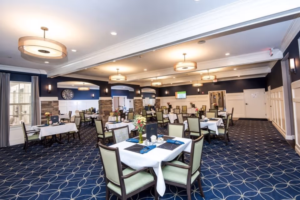 A spacious dining room with multiple tables covered in white tablecloths, each set with blue napkins, menus, and small flower arrangements. The room features blue patterned carpet, dark blue walls with white wainscoting, and modern ceiling lights. Large windows with curtains allow natural light to enter.