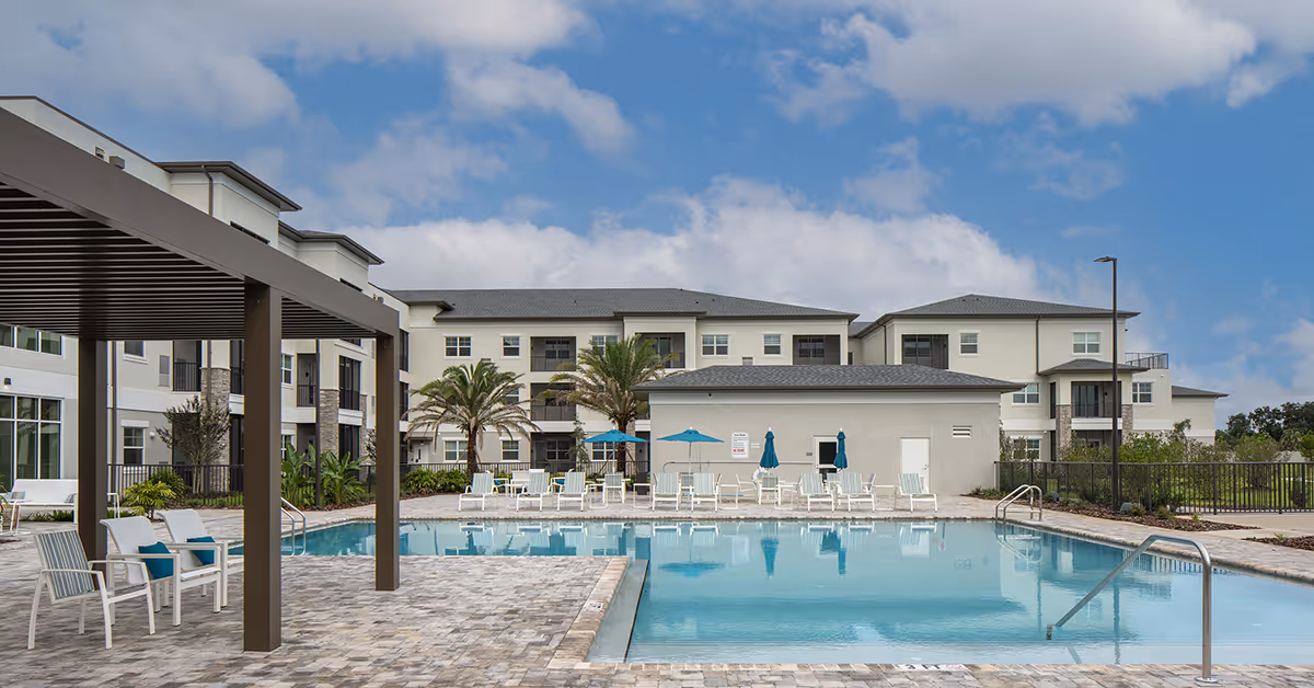Outdoor swimming pool area at a senior living facility with lounge chairs, umbrellas, palm trees, and a covered seating area. The building of the facility is visible in the background under a partly cloudy sky.