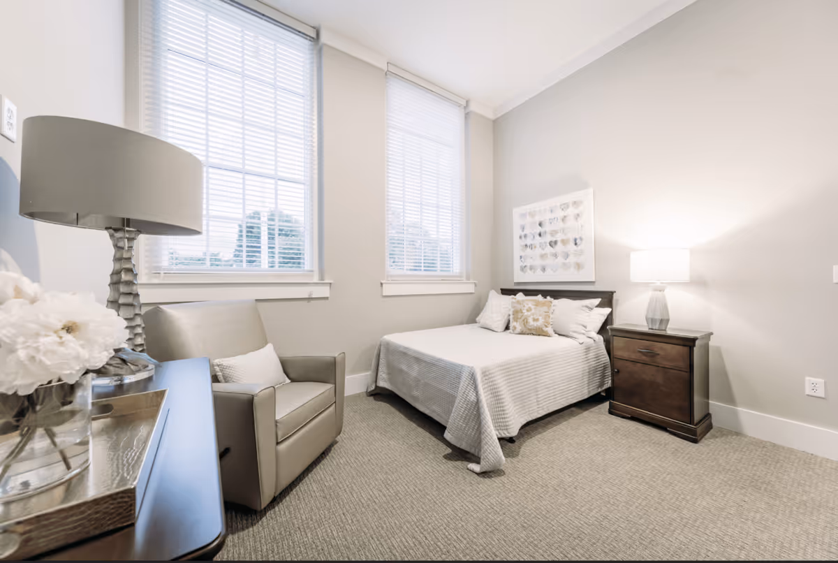 A bright and cozy bedroom with a bed covered in a light gray bedspread and decorative pillows. Next to the bed is a dark wooden nightstand with a modern lamp. There is a comfortable armchair beside a table with a lamp and a vase of white flowers. Two large windows with white blinds allow natural light to fill the room. The walls are painted a soft beige color, and a framed artwork featuring heart shapes hangs above the bed.