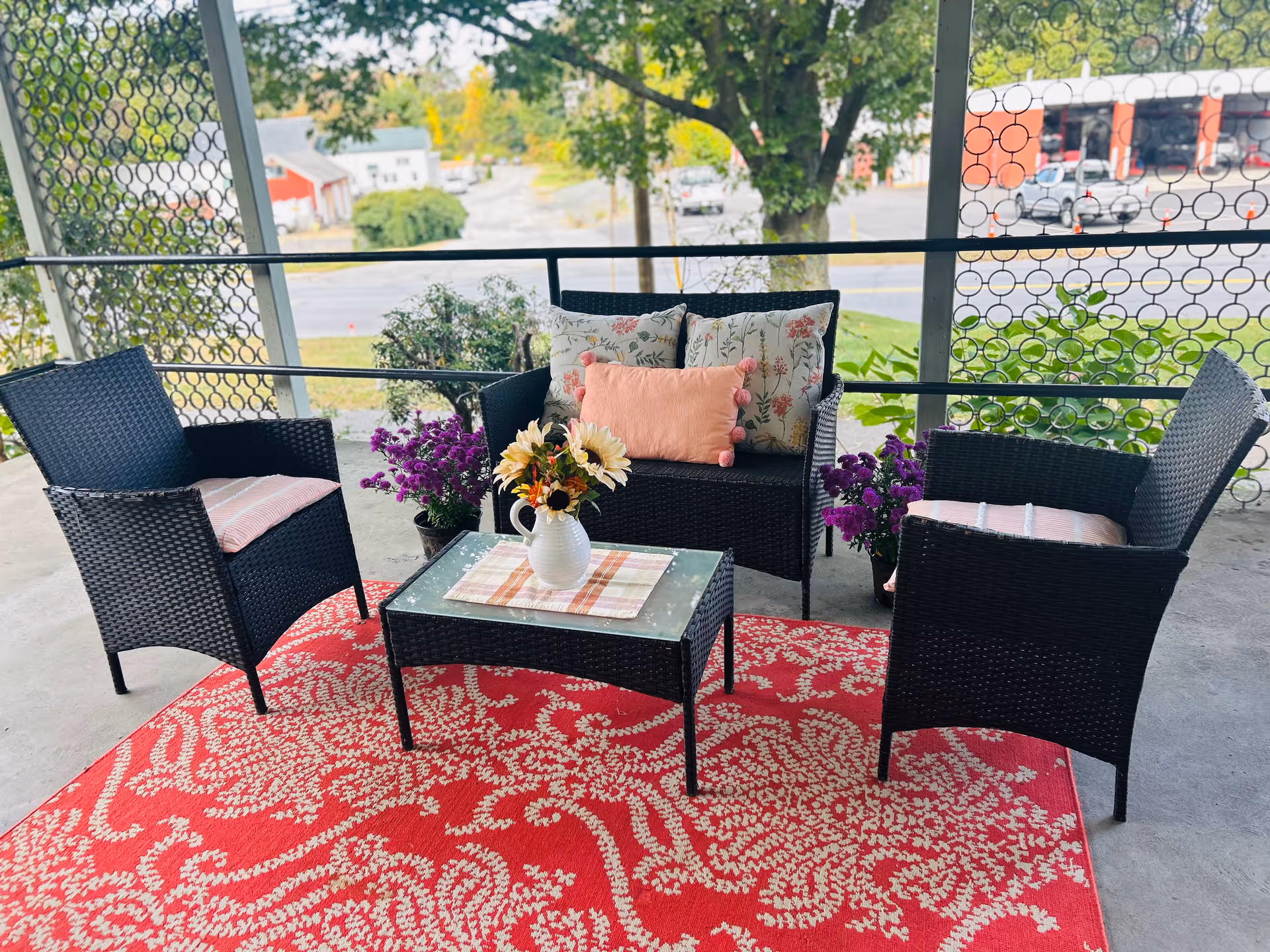Outdoor seating area with two black wicker chairs and a matching loveseat with floral and pink cushions, centered around a glass-top coffee table with a vase of sunflowers on a red patterned rug. The area is surrounded by a metal railing with circular designs, overlooking a street and trees in the background.