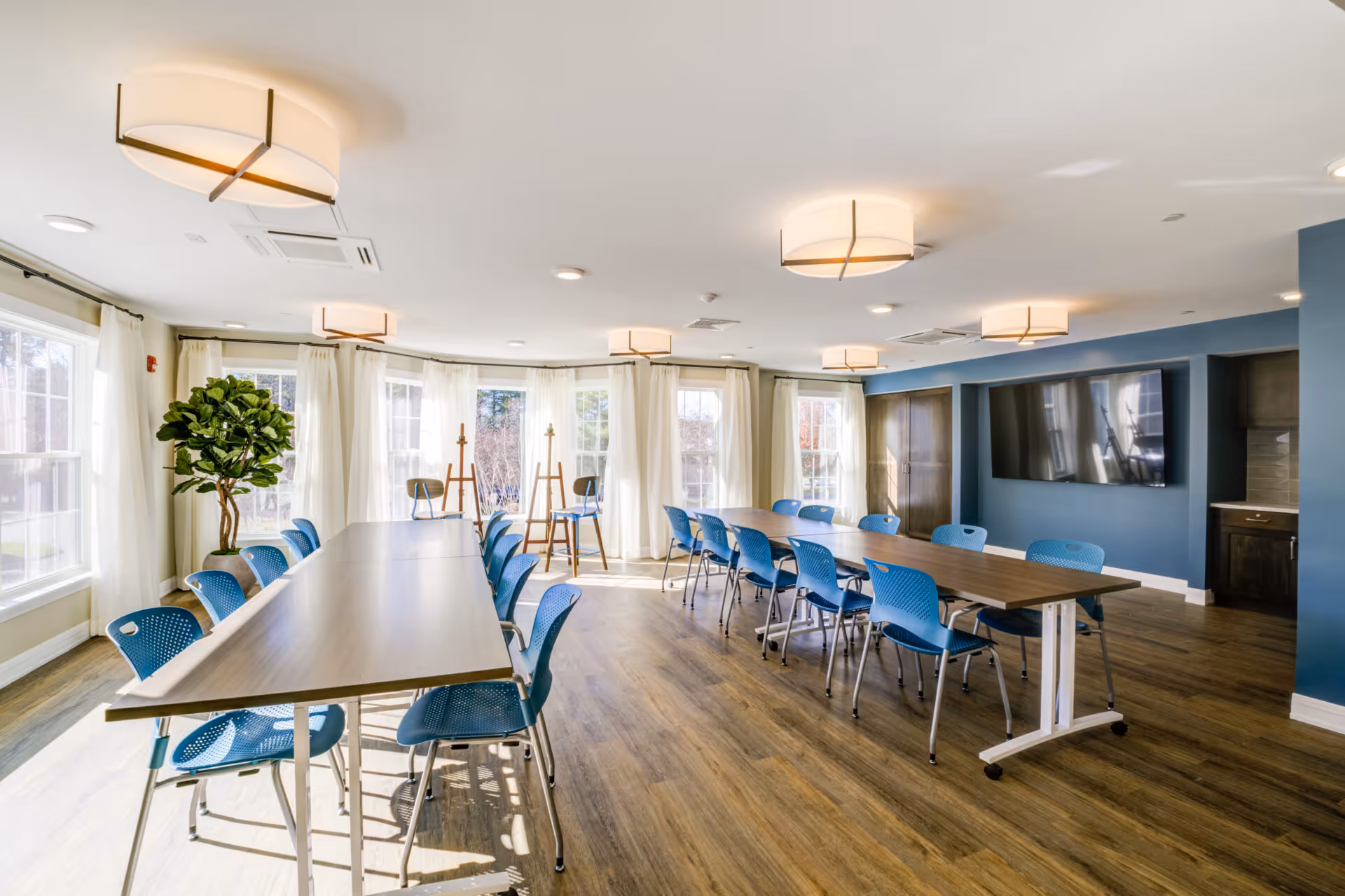 Bright multipurpose dining/activity room with long tables, blue chairs, large windows with curtains, a wall-mounted TV, and modern ceiling lights.