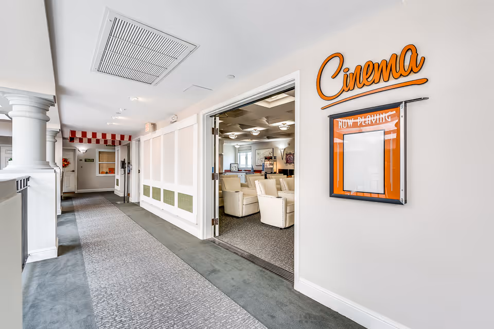 A bright hallway with carpeted flooring and white walls featuring decorative columns on the left side. On the right side, there is an open doorway leading into a room with beige armchairs arranged in rows, resembling a small cinema or theater. Above the doorway, an orange sign reads 'Cinema' with a 'Now Playing' display board below it.