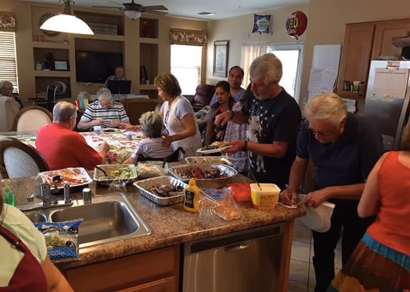 A group of elderly people and caregivers gathered in a kitchen and dining area. Some are seated at a dining table with food, while others are serving themselves from trays of food on the kitchen counter. The setting is warm and home-like with wooden cabinets, a sink, and a refrigerator visible.