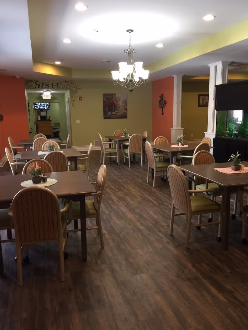 A dining room with multiple tables and chairs arranged neatly. Each table has a small plant centerpiece. The room has wood flooring, green and orange walls, a chandelier hanging from the ceiling, and a hallway labeled 'Suites 1-10' in the background.