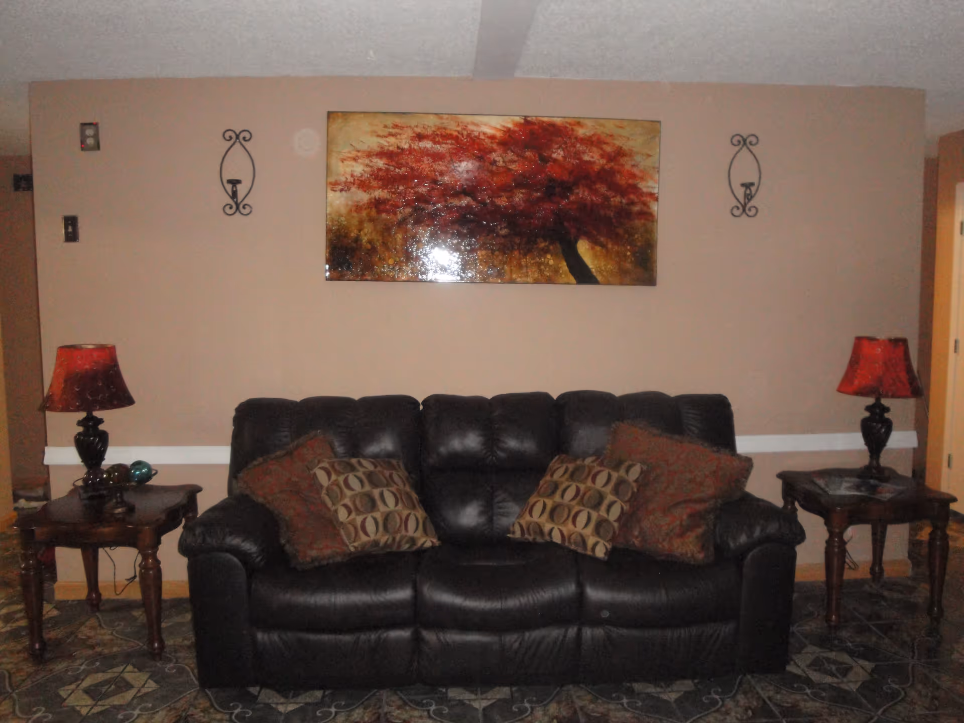 A cozy living room area featuring a black leather sofa with four decorative pillows. On either side of the sofa are wooden side tables with red lamps. Above the sofa hangs a painting of a tree with red leaves. The walls are painted beige and the floor has a patterned tile design.