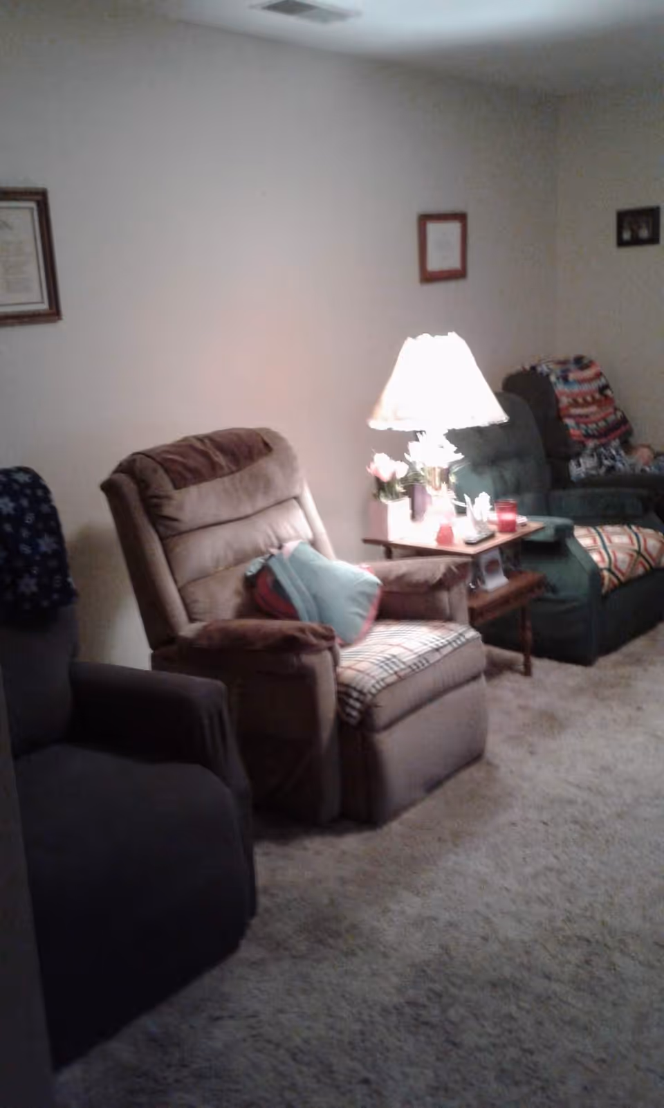 A cozy living room with three recliner chairs arranged along a wall. A side table with a lit lamp, flowers, and small decorative items is placed between two of the chairs. The room has carpeted flooring and framed pictures hanging on the wall.