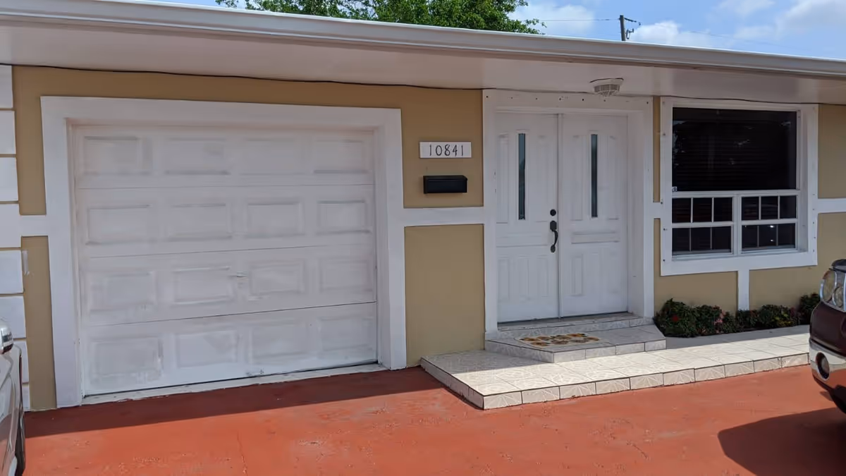 Front exterior view of a beige building with a white garage door, white double entrance doors, a window with white trim, and a small garden bed with flowers. The address number 10841 is displayed next to the door.