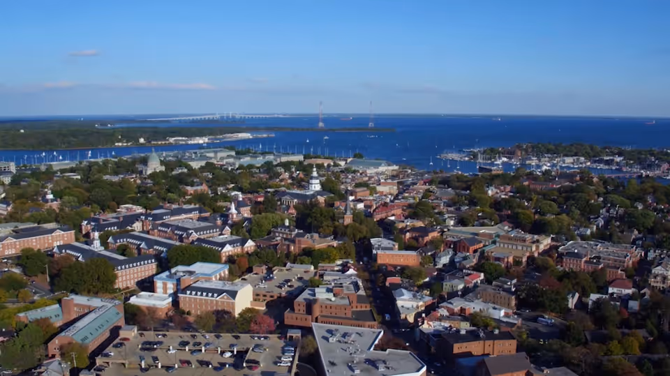 Aerial view of a coastal town with many brick buildings, streets, parked cars and a harbor with boats under a clear blue sky.