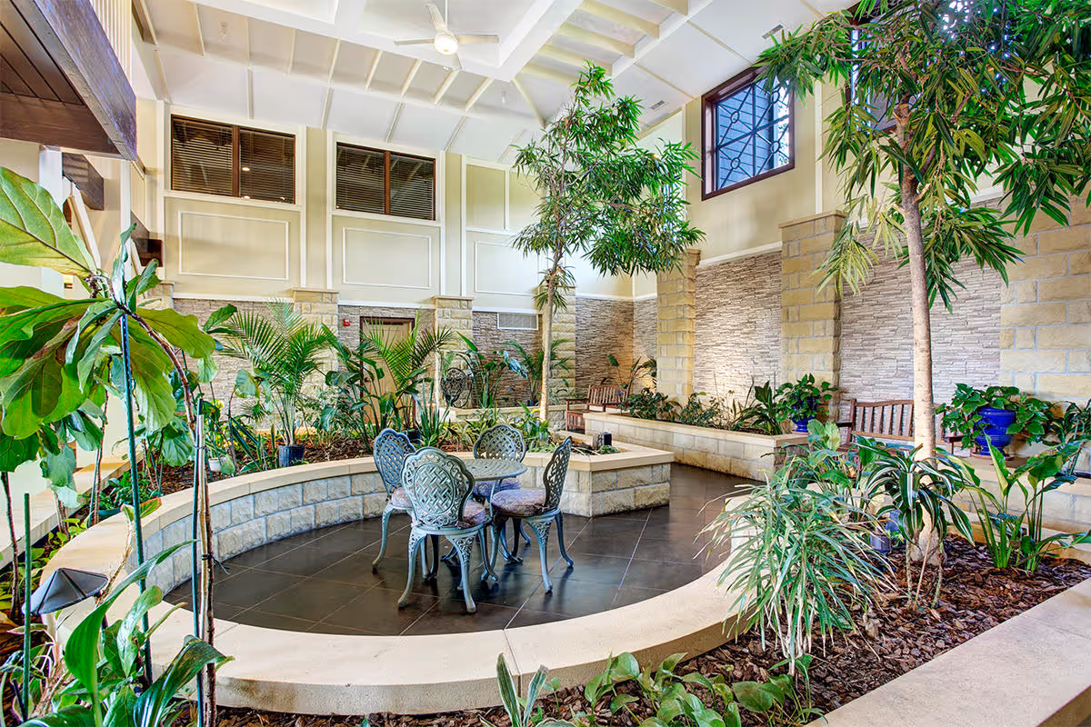 Indoor garden area with various green plants and small trees surrounding a circular seating area with a metal table and four chairs. The space has high ceilings with exposed beams, large windows, and stone walls with benches along the sides.