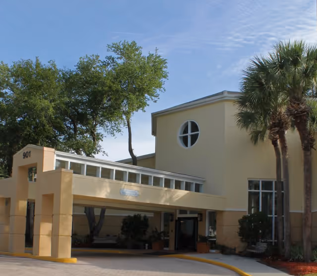 Exterior view of a beige senior living facility building with a covered entrance, large windows, palm trees, and other greenery under a clear blue sky.
