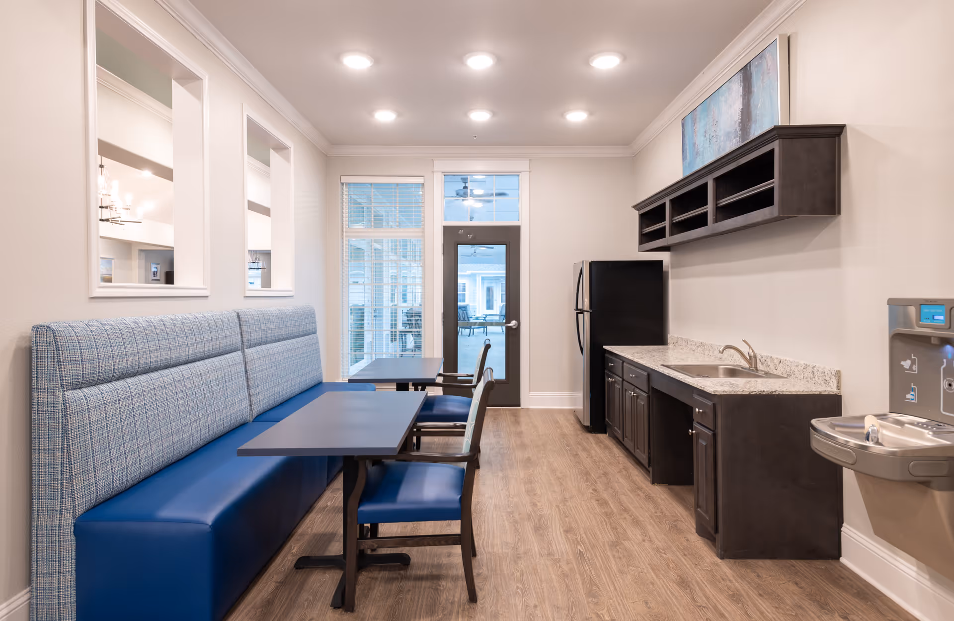 Small communal dining area with blue booth seating, tables and chairs alongside a kitchenette with dark cabinets, sink and refrigerator.