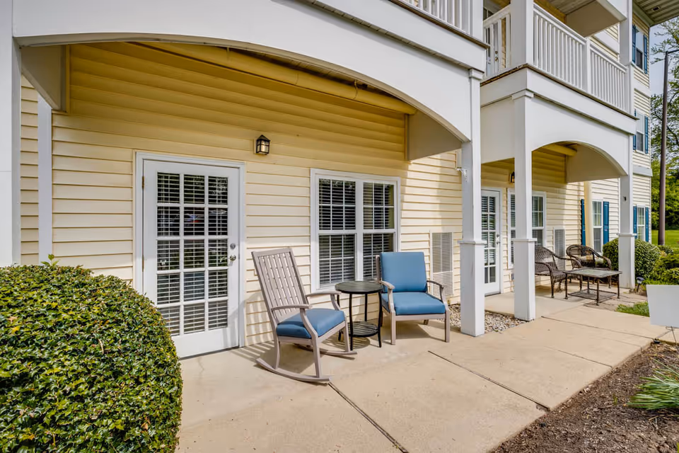 Outdoor patio area of a senior living facility with beige siding, white-framed glass doors and windows, blue cushioned chairs, a small round table, and neatly trimmed bushes along a concrete walkway.
