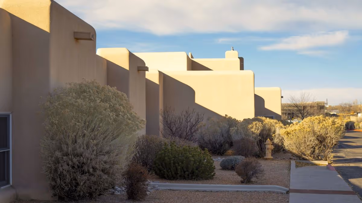 Exterior view of a building with smooth beige stucco walls and rounded edges, typical of Southwestern architecture. The building is surrounded by desert landscaping with various shrubs and bushes. A sidewalk and a fire hydrant are visible along the right side, under a partly cloudy sky.
