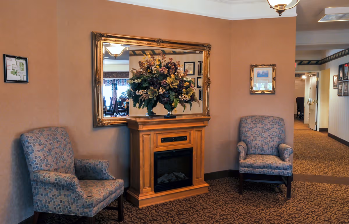 A cozy seating area with two patterned armchairs placed on either side of a wooden electric fireplace. Above the fireplace is a large ornate mirror reflecting a floral arrangement and part of a dining area. The walls are painted beige, and the floor is carpeted with a patterned design. There are framed pictures and a floor plan on the walls, and a hallway is visible in the background.