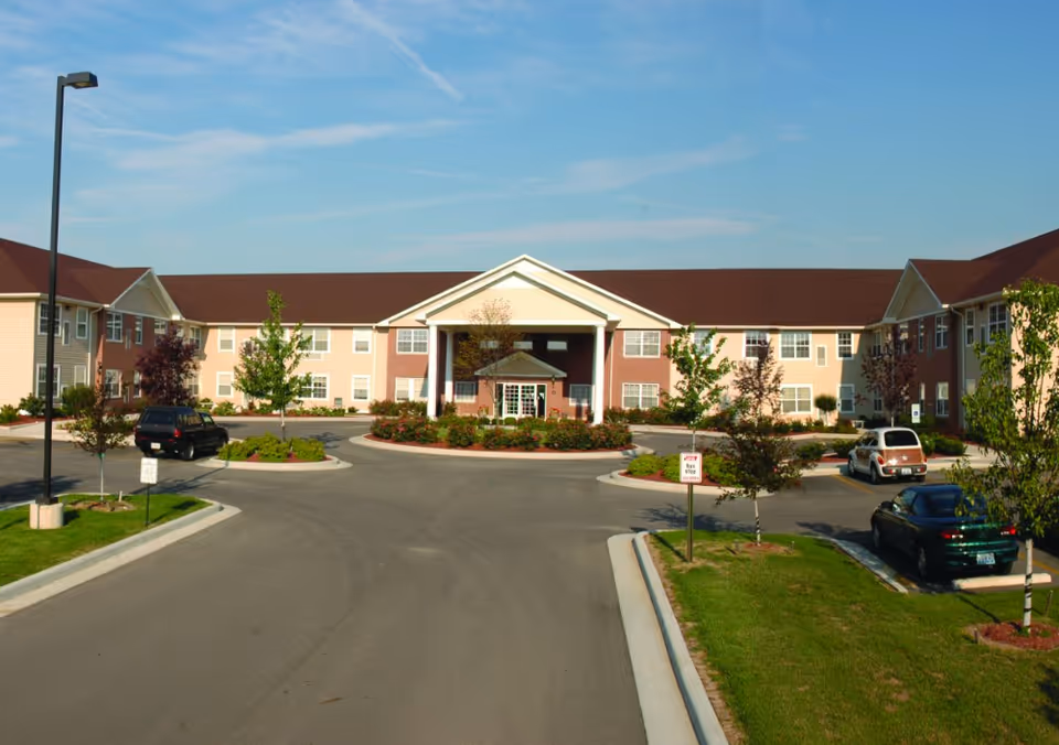 Front exterior view of a two-story senior living facility building with a central entrance featuring a covered portico, surrounded by landscaped greenery and a circular driveway with parked cars under a clear blue sky.