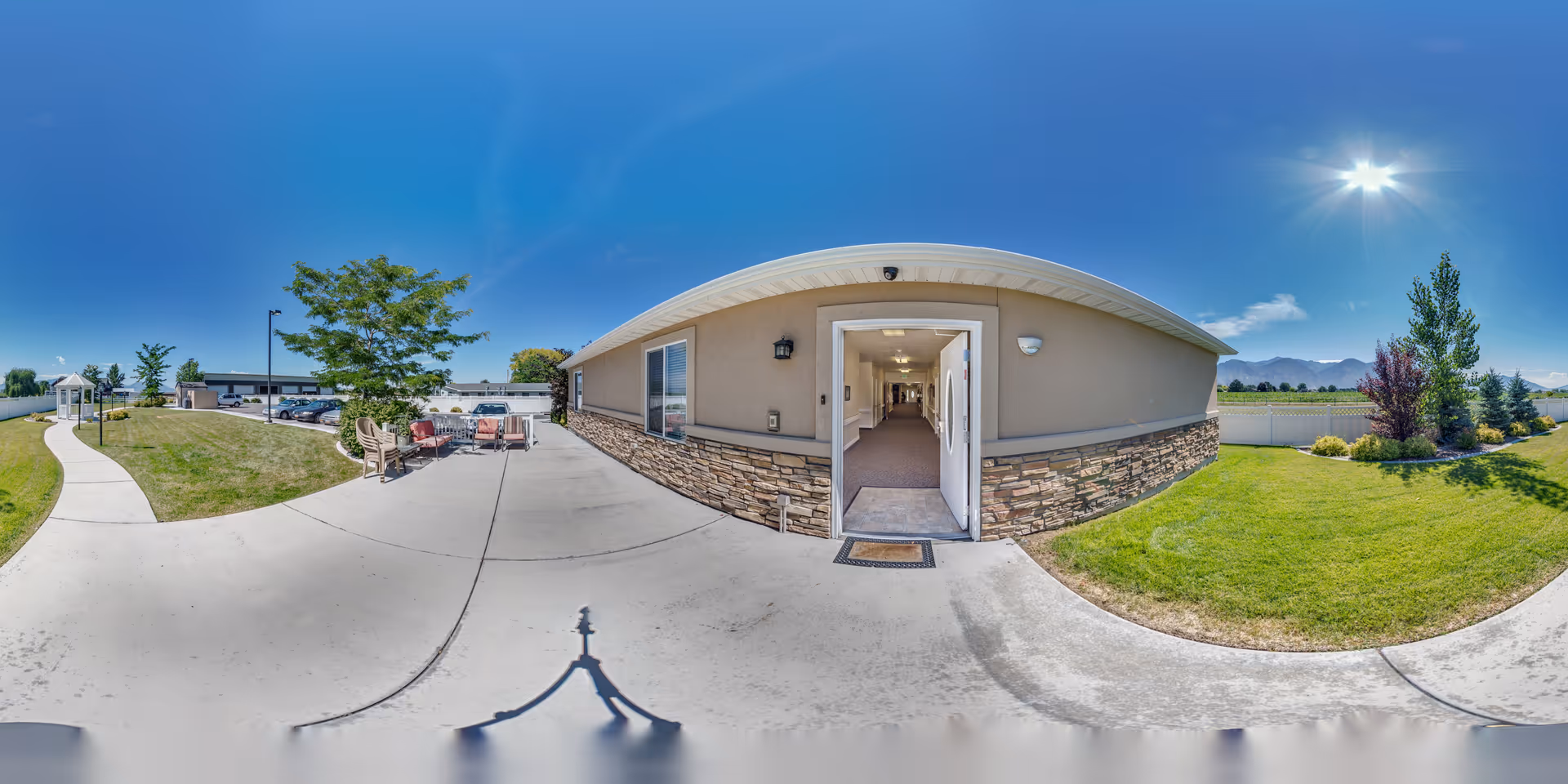 Outdoor view of BeeHive Homes of Provo showing a curved concrete walkway, green grassy areas with trees and shrubs, outdoor seating with chairs and tables, and the entrance to a building with an open door leading to a hallway. The sky is clear and sunny with mountains visible in the background.