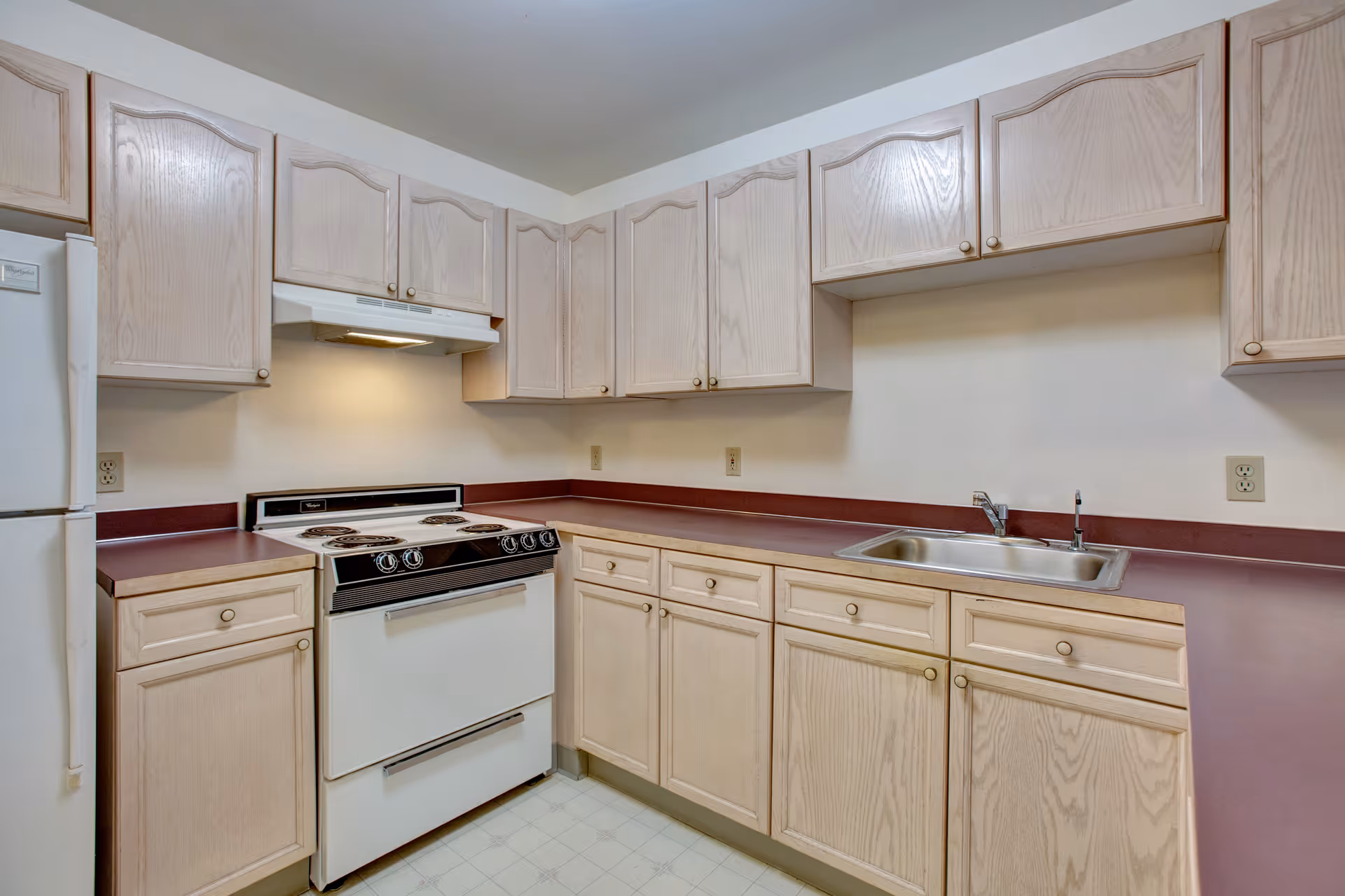 A kitchen with light wood cabinets, a white electric stove with four burners, a white refrigerator, a stainless steel sink, and maroon countertops. The walls are light-colored and there is a white range hood above the stove.