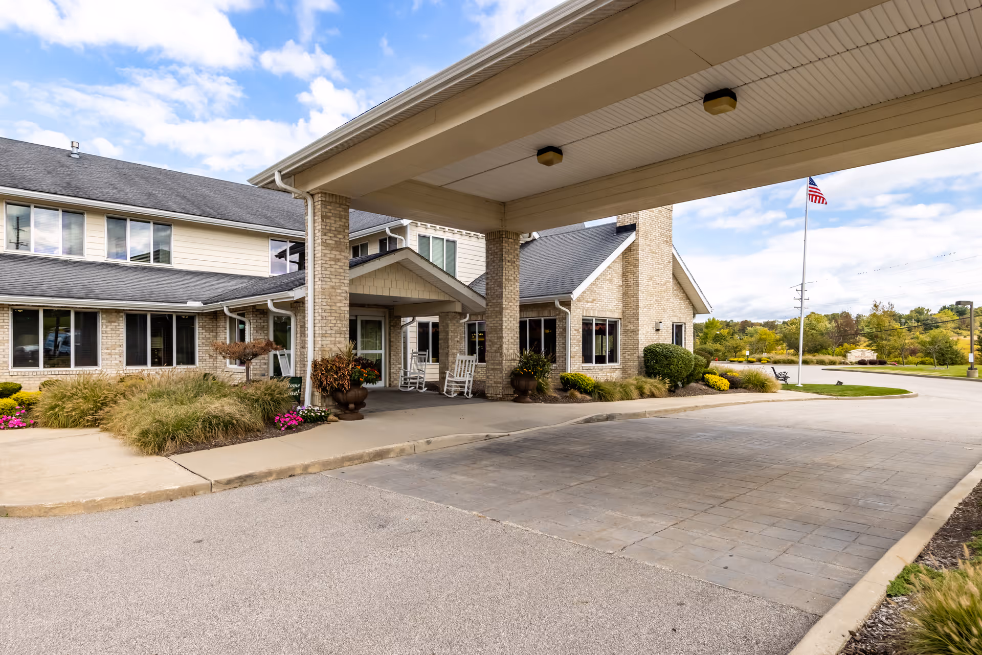Entrance of a senior living facility with a covered drop-off area supported by brick columns, rocking chairs on the porch, landscaped bushes and flowers, and an American flag on a flagpole in the background under a partly cloudy sky.