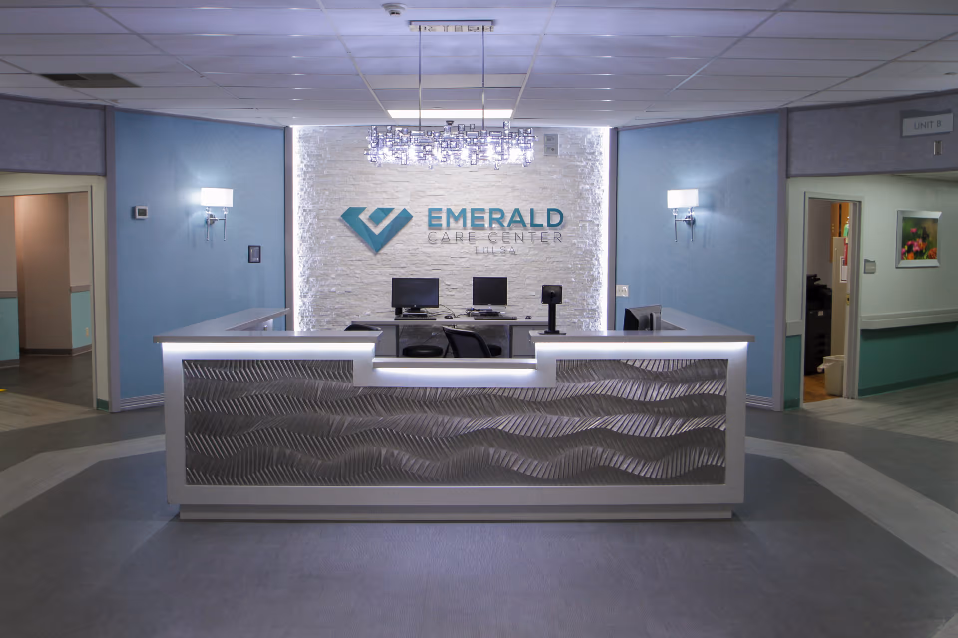 Reception desk area inside Emerald Care Center Tulsa with a modern front desk featuring a textured wave pattern, two computer monitors, and a chandelier overhead. The wall behind the desk has the Emerald Care Center Tulsa logo mounted on a white textured surface. The surrounding walls are painted light blue with wall-mounted lights, and doorways lead to other parts of the facility.