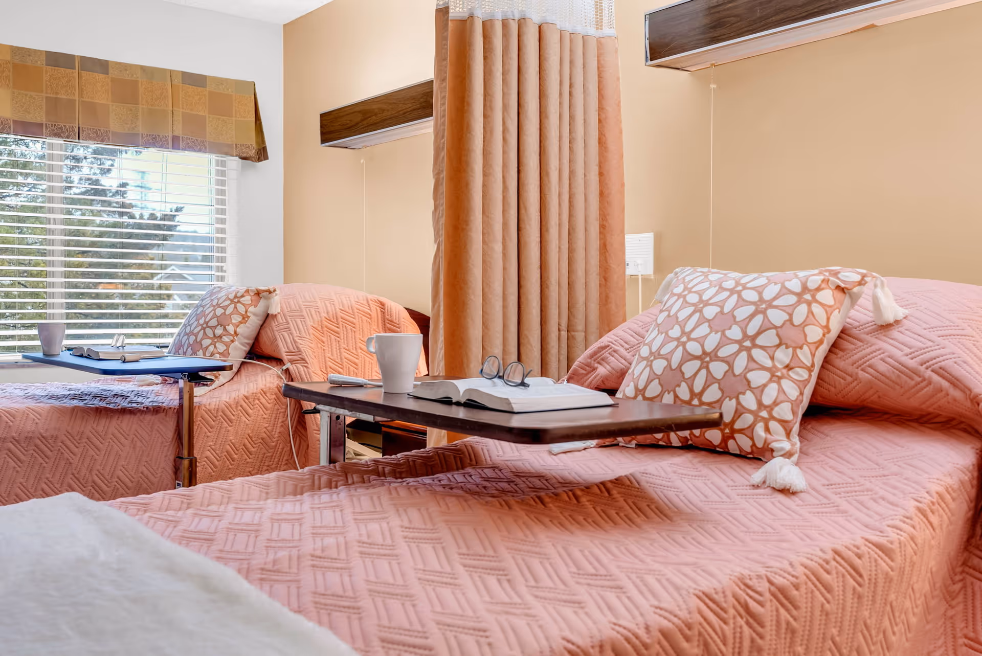Shared senior care bedroom with two twin beds dressed in pink quilts, overbed trays, a privacy curtain, and a window with blinds.