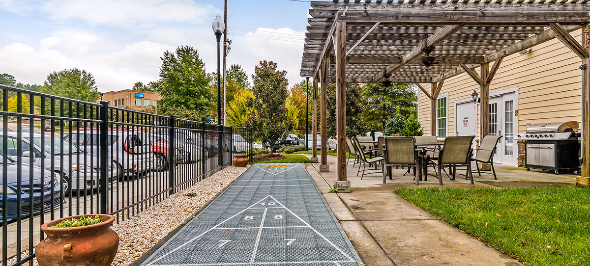 Outdoor patio area with a shuffleboard court leading to a pergola-covered seating area and a grill beside a parking lot.