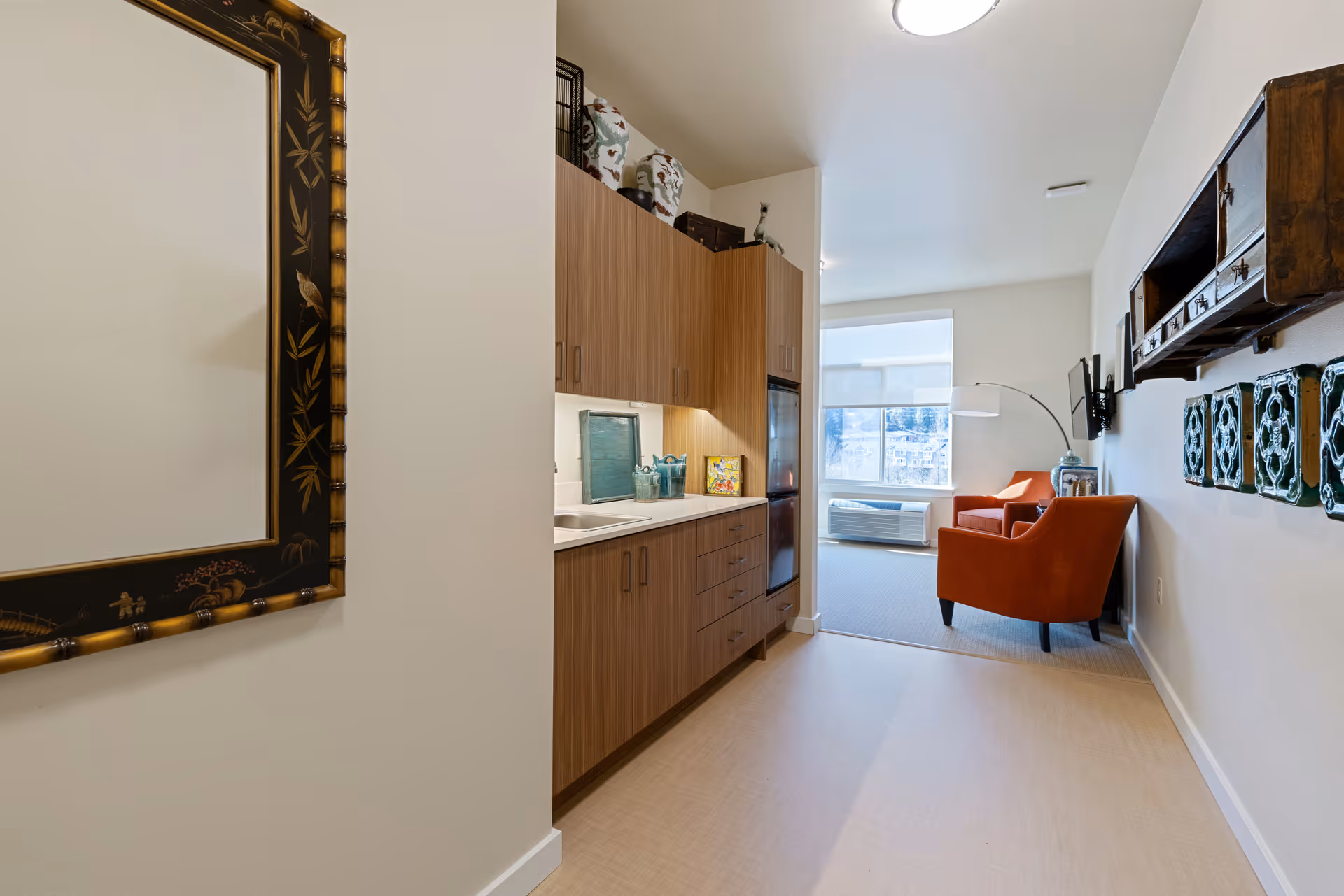 Interior view of a senior living facility showing a hallway with wooden cabinets and a countertop on the left, leading to a small living area with two orange armchairs, a wall-mounted TV, and a large window with a white blind. Decorative items and artwork are displayed on the walls.