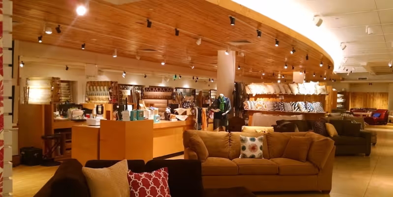 Open indoor showroom-style living area with sofas, pillows, wooden displays and warm overhead lighting.