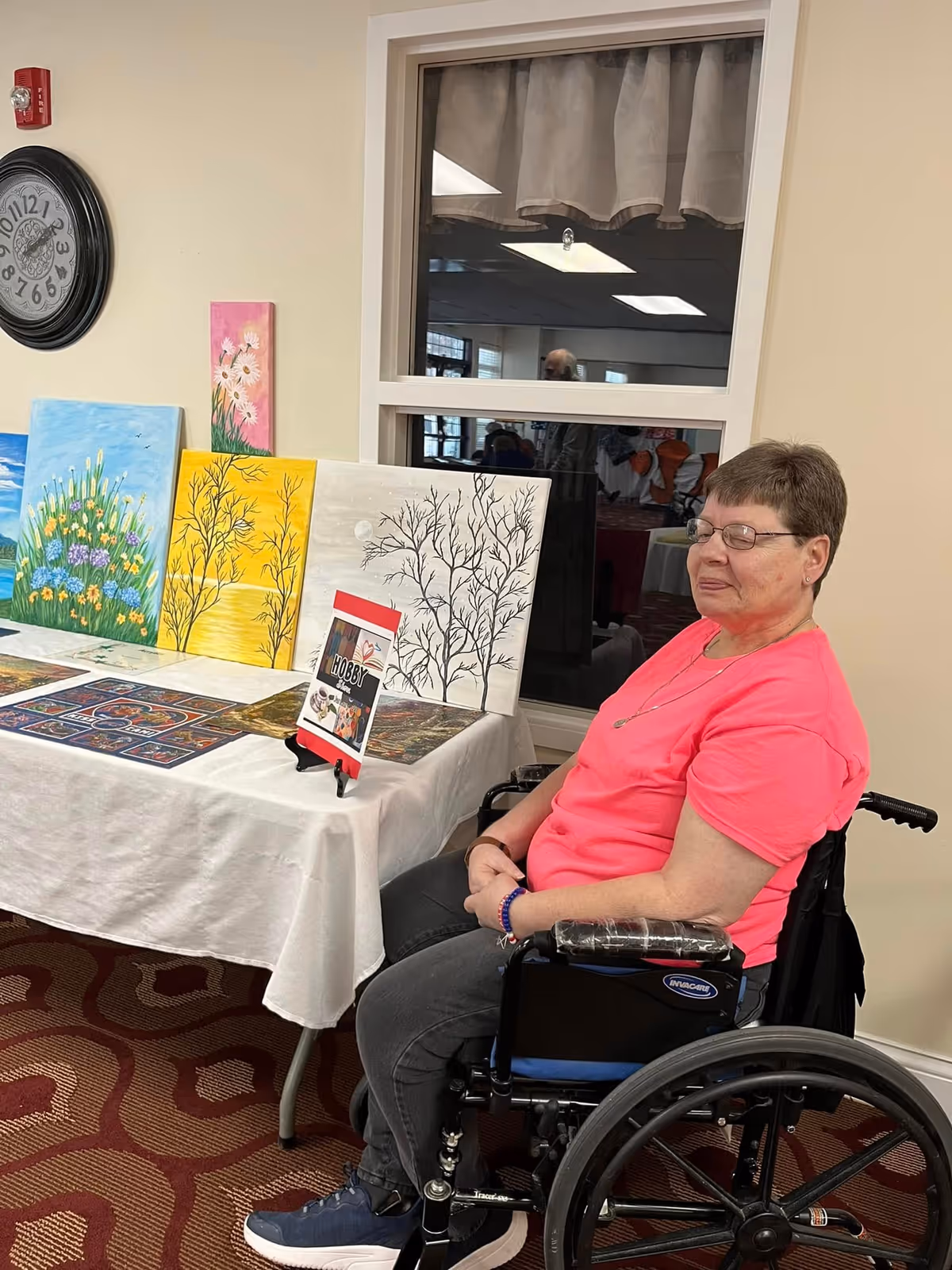 A woman in a wheelchair wearing a bright pink shirt and glasses sits next to a table displaying various colorful paintings and artwork. The setting appears to be indoors with a window and a clock on the wall behind her.