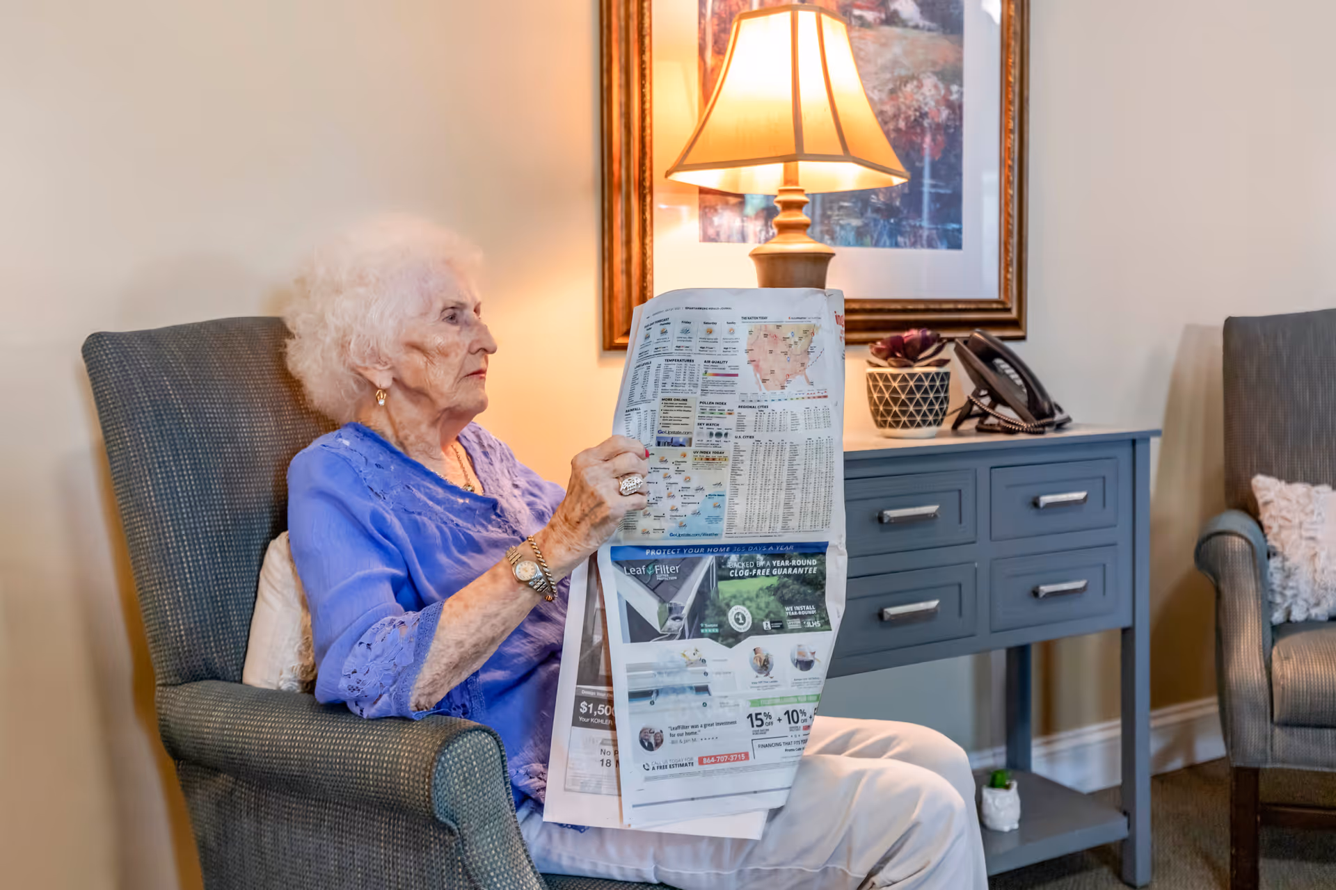 An elderly woman with white hair wearing a blue blouse and white pants is sitting in a gray armchair reading a newspaper. Behind her is a blue side table with drawers, a table lamp, a potted plant, and a telephone. A framed artwork hangs on the wall above the table.