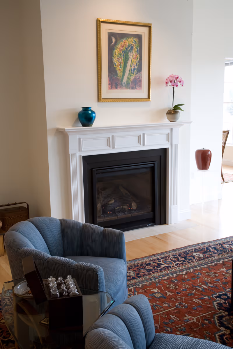 Cozy living room with a white fireplace mantel, framed artwork above, blue upholstered chairs, and a patterned area rug.